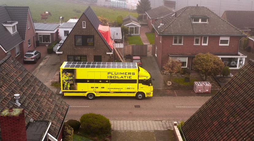 Afbeelding van een straatbeeld. Voor een huis, waar aan het dak gewerkt wordt, staat een gele vrachtwagen van Pluimers Isolatie.